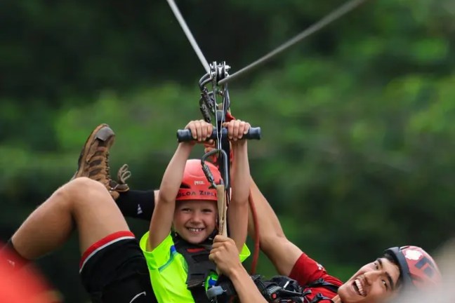 Child and adult zip-lining together, both smiling, with green foliage background.